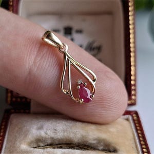 May include: A gold pendant featuring a pear-shaped ruby gemstone and a tiny diamond. The pendant is held in a hand, highlighting its intricate design and vivid red hue. The pendant is in a red jewellery box.