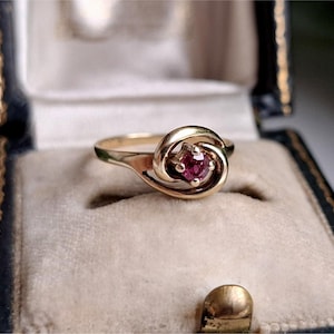 Könnte beinhalten: Ein Goldring mit einem runden, facettierten, dunkelrosa Edelstein, der in ein geschwungenes Band im Bypass-Stil eingefasst ist. Das Metall hat eine polierte Oberfläche und der Stein wird von vier Krappen in der Mitte des Designs gehalten.