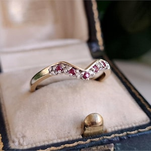 Op de afbeelding: Een gouden ring in chevronvorm met een rij kleine, rond geslepen rode robijnen en heldere accentstenen gezet langs de V-vormige band. De ring wordt gepresenteerd in een donkerkleurig, scharnierend juwelendoosje met een crèmekleurige fluwelen voering.
