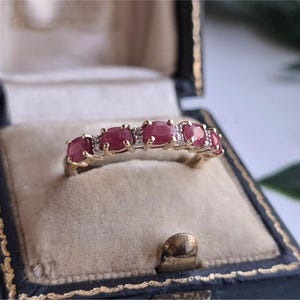 May include: A gold ring featuring five oval-cut red rubies set in a row, accented by small clear stones between each ruby. The ring is displayed inside a dark blue velvet-lined jewellery box with a cream-coloured interior cushion.