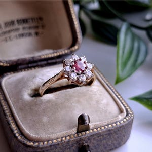 Puede incluir: Un anillo de oro vintage con una gema roja central rodeada de piedras blancas más pequeñas, dispuestas en un diseño floral. El anillo se exhibe en un joyero antiguo abierto, con interior de terciopelo. La caja es marrón con ribetes dorados.