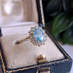 Könnte beinhalten: Ein Goldring mit einem zentralen, oval geschliffenen hellblauen Edelstein, der von einem Kranz aus kleineren, runden, durchscheinenden weißen Steinen umgeben ist. Der Ring hat eine dünne Goldschiene und liegt in einem dunkelblauen Schmuckkästchen.