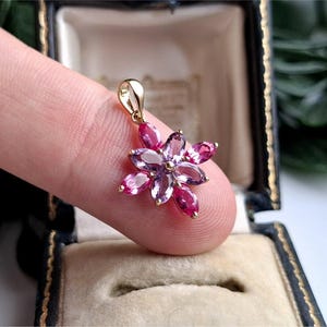May include: A gold pendant featuring a flower-shaped arrangement of marquise-cut gemstones. The center and inner petals are light purple, while the outer petals are a vibrant magenta pink. The pendant includes a small gold bail at the top.
