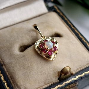 Puede incluir: Un colgante de oro en forma de corazón con un ópalo redondo central rodeado por seis pequeños rubíes rojos. La superficie dorada del corazón está decorada con un patrón de estrella texturizado y el colgante incluye una pequeña argolla de oro.