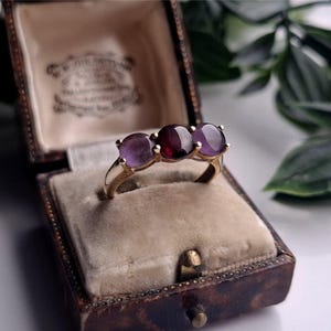May include: A gold ring featuring a central red garnet and two purple amethyst gemstones. The ring is presented in a vintage brown leather jewellery box with a beige velvet lining. The ring's setting is gold.
