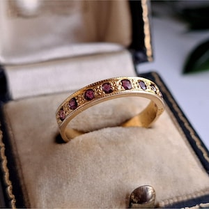 May include: A gold ring with a band of small, round, dark red gemstones. The ring is set in a vintage jewelry box with a blue exterior and a cream-colored interior. The ring's band has a decorative pattern.