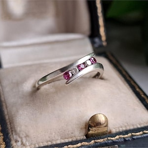 May include: A silver-toned bypass ring featuring a row of three round-cut pinkish-red stones alternating with two small clear stones set within a curved, polished metal band that overlaps slightly at the centre of the design.