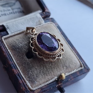 May include: A gold pendant with a purple oval gemstone set in a decorative filigree frame. The pendant is displayed in a velvet-lined jewelry box.