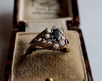 May include: A gold ring with a black oval gemstone surrounded by smaller white gemstones. The ring is in a brown velvet ring box.
