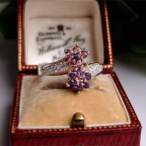 Op de afbeelding: Een gouden ring met een bloemmotief, bezet met roze en paarse edelstenen. De ring is gezet met kleine witte diamanten en wordt gepresenteerd in een rode sieradendoos.