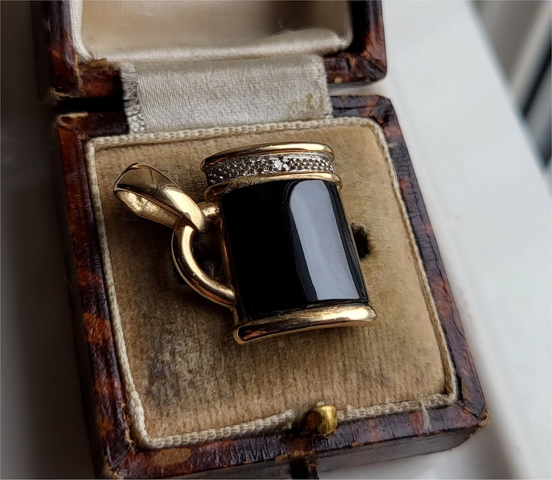 May include: A gold-tone pendant in the shape of a beer stein with a black onyx stone and a row of clear cubic zirconia stones around the top.