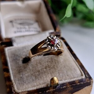 May include: A gold ring featuring a red gemstone surrounded by smaller white gemstones. The ring is displayed in a velvet-lined box.