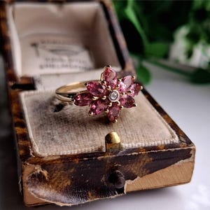 Könnte beinhalten: Ein goldener Ring mit einem rosafarbenen Blumendesign. Die Blume besteht aus rosafarbenen Edelsteinen und hat einen einzelnen weißen Edelstein in der Mitte.