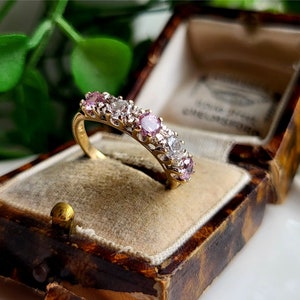 Puede incluir: Un anillo de banda de oro con una fila de piedras preciosas rosadas y blancas engastadas en un diseño delicado e intrincado. El anillo se muestra en una caja de joyería forrada de terciopelo marrón.