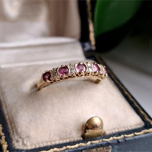 May include: A gold band ring featuring a row of five round-cut pink gemstones alternating with small clear accent stones. The ring is set in a dark blue jewellery box with a cream-coloured velvet interior lining, showcasing the metal and stone details.