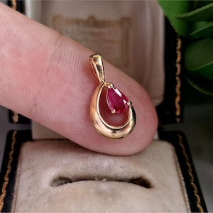 May include: A gold pendant featuring a teardrop-shaped red gemstone set within a smooth, polished gold open-loop frame. The pendant has a small integrated bail at the top for attachment to a chain, showcasing a minimalist design with a vibrant stone.