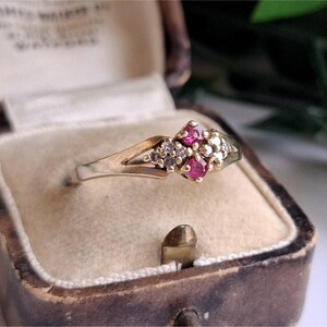 May include: A gold ring featuring two small round red rubies and a cluster of tiny clear white diamonds set in a bypass style band. The ring is positioned inside a vintage-style brown jewellery box with a cream-coloured velvet interior lining.
