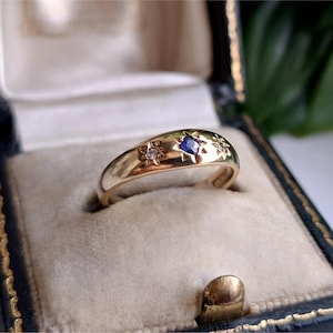 Op de afbeelding: Een gouden ring met een centrale blauwe edelsteen en twee kleinere heldere stenen, elk gezet in een stervormige gravure op het oppervlak van het metaal. De ring heeft een gepolijste afwerking en een afgerond profiel.