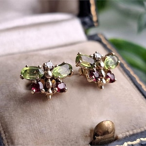 May include: A pair of gold stud earrings shaped like insects, featuring oval-cut light green peridot wings, round-cut deep red garnet bodies, and a small clear white diamond accent at the head of each piece, presented in a dark blue jewelry box.