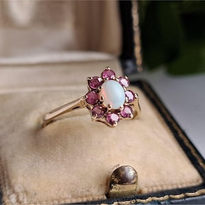 May include: A gold ring with an oval opal centre stone, encircled by small, round, pink gemstones. The ring is set in a gold band and presented in a vintage jewellery box.