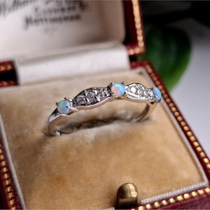 May include: A silver ring with a curved design, featuring small opals and clear gemstones. The ring is displayed inside a vintage red jewelry box with a gold clasp and a beige interior.