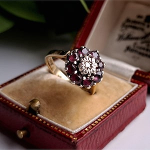 May include: A gold ring with a cluster of dark red gemstones surrounding a central silver-colored floral design. The ring is displayed in a red jewelry box with a tan interior.