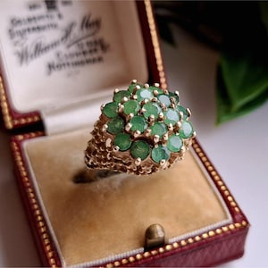 May include: A gold ring with a cluster of round, green emeralds. The ring is set in a textured gold band and is displayed in a red and tan jewelry box.