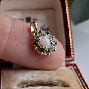 Puede incluir: Colgante de oro con una piedra de ópalo ovalada en el centro, rodeada de pequeñas piedras verdes. El colgante está engastado en un marco dorado y se presenta en un joyero vintage.