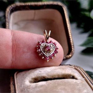 May include: A heart-shaped pendant crafted from yellow gold, featuring a central round-cut red ruby surrounded by a halo of smaller round-cut red rubies and tiny clear accent stones. The pendant includes a gold bail at the top for attachment.