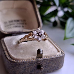 May include: A vintage gold ring with a cluster of diamonds and a central dark gemstone, presented in a worn, antique jewelry box. The ring's design features a floral arrangement of small, round diamonds. The box is gray with a velvet interior.