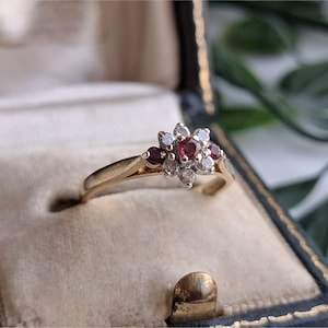 May include: A gold ring featuring a cluster setting with a central red gemstone surrounded by smaller clear stones and two additional red gemstones on the sides. The ring is displayed in a dark blue velvet-lined jewellery box with a cream interior.