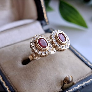 May include: A pair of gold stud earrings featuring a central oval-cut red ruby surrounded by a halo of small white diamonds. The earrings have a scalloped edge design and are set in a yellow gold metal frame with a polished finish.