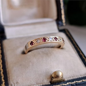 Puede incluir: Un anillo de banda de oro con una fila alterna de rubíes rojos y diamantes blancos transparentes de talla redonda engastados en el metal. El anillo está colocado dentro de un joyero oscuro con un forro interior acolchado de color claro.