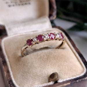 May include: A gold band ring featuring a row of three round red rubies alternating with two small clear diamonds. The ring is set in a beige velvet-lined jewelry box with a dark brown exterior, showcasing a classic multi-stone design.