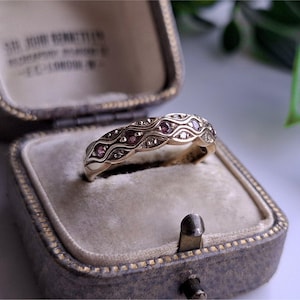 May include: A gold ring with a wavy design, set with small pink and white gemstones. The ring is displayed inside a vintage gray jewelry box with a velvet interior. The box is open, showcasing the ring's intricate details.