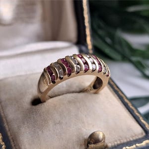 May include: A gold band ring featuring a channel setting with alternating round-cut red rubies and clear white diamonds. The ring has a curved, arched profile with metal bars separating each gemstone, creating a structured, multi-stone design.