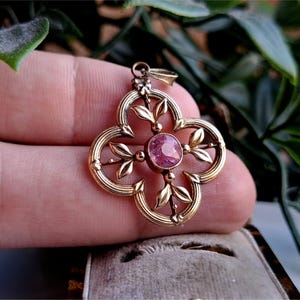 May include: A gold-toned pendant with a four-leaf clover design, featuring a central pink gemstone. The pendant has delicate leaf and bead details, and a small loop for a chain.
