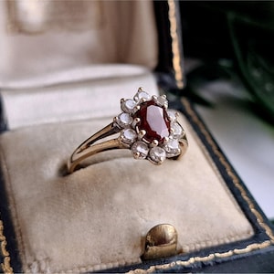 Può includere: Un anello in oro con una gemma centrale rosso scuro dal taglio ovale, circondata da un alone di piccole pietre bianche rotonde e trasparenti. Il gambo dell'anello è diviso sulle spalle ed è presentato in un cofanetto foderato in velluto blu.