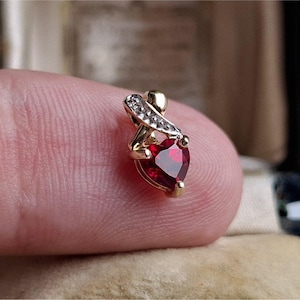 Puede incluir: Colgante de oro con una piedra preciosa roja oscura en forma de corazón y facetada. El colgante tiene una pequeña parte superior curvada con un acento con diamantes. La piedra preciosa mide aproximadamente 1,3 cm de largo. Joyería.