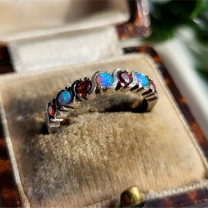 May include: A silver band ring with alternating red and blue gemstones. The ring is set in a velvet box.