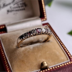 May include: A gold ring with a row of gemstones, including red and clear stones, rests in a vintage red and tan jewelry box. The ring is centered in the box, with the box open. The box has a gold clasp and a label inside.