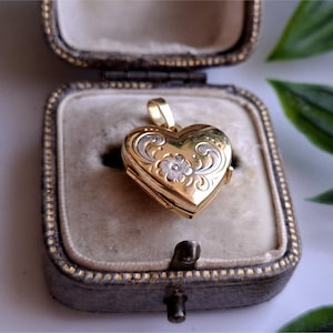 Peut inclure: Un médaillon doré en forme de cœur avec gravure florale et bélière. Le médaillon est présenté ouvert dans un écrin à bijoux gris vintage avec un intérieur en velours. Le médaillon a une fermeture à charnière et un design détaillé.