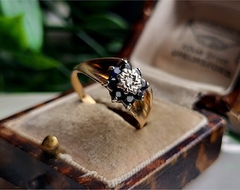 May include: A gold ring with a flower-shaped setting featuring a cluster of blue sapphires and a single diamond in the center.