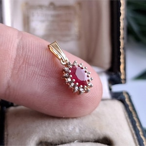 Peut inclure: Un pendentif en or orné d'un rubis rouge central de taille ovale, entouré d'un halo de petits diamants blancs. Le pendentif possède une bélière en or poli sur le dessus pour l'attacher à une chaîne, présentant un design classique à griffes.