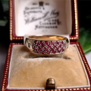 Puede incluir: Un anillo de oro con una banda de pequeñas gemas rojas vibrantes. El anillo está engastado en un joyero rojo vintage con ribetes dorados y un interior color crema. La caja tiene texto en el interior de la tapa.