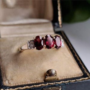 Può includere: Un anello d'oro con tre pietre preziose ovali rosso scuro e piccole pietre di accento. L'anello è presentato in un portagioie nero vintage con chiusura dorata e interno beige.