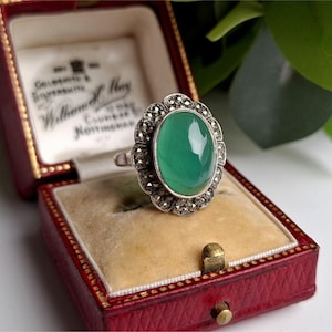 Könnte beinhalten: Ein silberner Vintage-Ring mit einem großen ovalen grünen Edelstein, umgeben von kleineren Steinen. Der Ring wird in einer offenen roten Schmuckschatulle mit cremefarbenem Innenraum präsentiert. Die Schatulle ist mit Text geprägt.