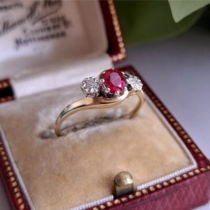Może przedstawiać: Złoty pierścionek z dużym owalnym rubinem pośrodku i dwoma mniejszymi diamentami. Pierścionek jest prezentowany w czerwonym pudełku na biżuterię z kremowym wnętrzem. Projekt jest w stylu vintage.