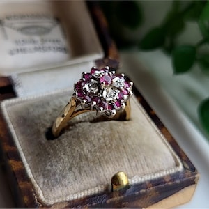 Può includere: Un anello d'oro con un gruppo di rubini rossi e diamanti bianchi incastonati a forma di fiore. L'anello è in una scatola di gioielli foderata in velluto.