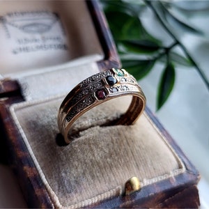 以下が含まれることがあります： 小さなダイヤモンドと宝石がセットされた、3つのバンドを持つゴールドリング。リングは、ベルベットの内装が施されたヴィンテージのジュエリーボックスに展示されています。宝石は赤、緑、黒です。